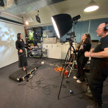 Woman standing on a platform in a studio, being filmed by a camera crew with lights and equipment.